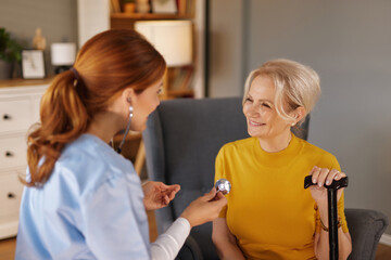 Fototapeta premium A young woman with red hair, wearing a blue uniform, uses a stethoscope on a smiling senior woman with short blond hair in a yellow shirt. The senior woman is sitting in a chair with a cane.