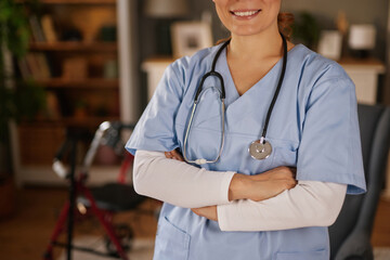 A smiling nurse stands with crossed arms, wearing scrubs and a stethoscope in a home setting. A...