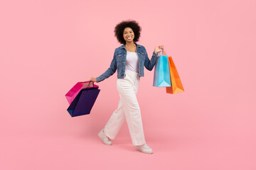 Happy young black woman walking confidently with colorful shopping bags, smiling african american female enjoying making purchases, posing on vibrant pink studio background, copy space