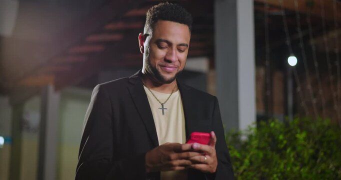 Hispanic man of African descent smiling at red smart phone while outdoors at night, joyful reaction to message under string lights in urban garden setting