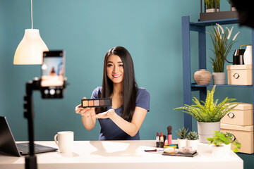 Friendly asian vlogger presenting an eyeshadow palette towards her cellphone camera, recording a video. She demonstrates makeup techniques, holding a product while filming for an online tutorial.
