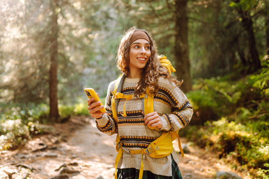 Smiling female hiker with bright yellow backpack using phone on hiking trail. Beautiful woman enjoying adventure and hiking in mountains. Travel and blogging concept.