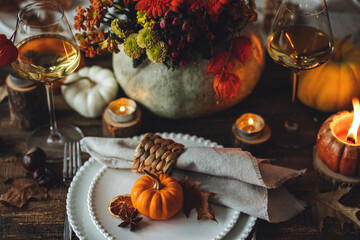 Elegant seasonal fall table setting with floral composition in a pumpkin vase as a centrepiece. White wine, burning candles, romantic cozy atmosphere of family dinner, private autumn wedding