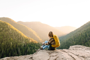 Fototapeta premium Beautiful woman hiker feels freedom standing on rocky ledge enjoying sunset in mountains. Young female hiker looks at mountain landscape on cliff. Hiking, adventure and nature concept.