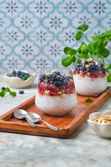 Chia pudding with nut granola, honeysuckle berry puree and whole honeysuckle  berries in transparent glasses on grey concrete background. No bake desserts, desserts in glasses.