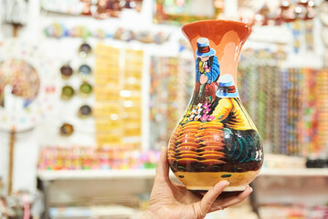 Female hand holding a handcrafted ceramic vase painted with a colorful traditional scene, in...