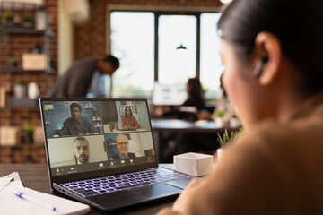 Selective focus on laptop displaying group of coworkers on a videoconference call with a businesswoman wearing earphones. Female manager sitting at desk, attending an online meeting with clients.