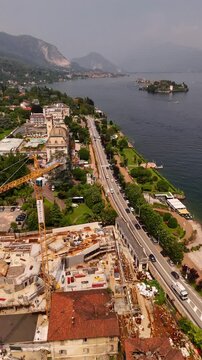 Drone video of a tall construction crane towering above a lakeside town with red rooftops and panoramic views of Lake Maggiore, Italy. Urban development meets alpine and lakefront scenery