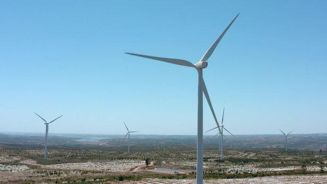 Multiple wind turbines are rotating against a clear blue sky, overlooking an agricultural landscape with fields and a body of water in the distance