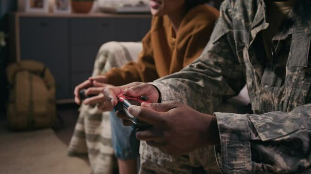Medium midsection shot of hands of anonymous black male soldier in khaki uniform and young son sitting on couch together, playing and competing in video game, boy grabbing fathers controller