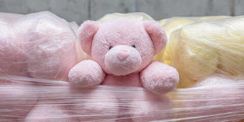 Pink teddy bear sits on plastic wrapped bundles. Softness, innocence, childhood, comfort, plush, gentle, cozy, pastel, playful, toy nostalgia
