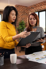 Asian businesswomen collaborate on strategy planning, comparing ideas with a clipboard and tablet in a modern workspace. Two female colleagues stand in office, reviewing each others project notes.