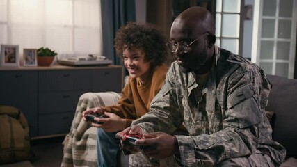 Medium shot of cheerful, laughing black male army officer in camouflage uniform and teenage biracial son sitting on couch at home, playing console video game together, competing, laughing with joy - Powered by Adobe