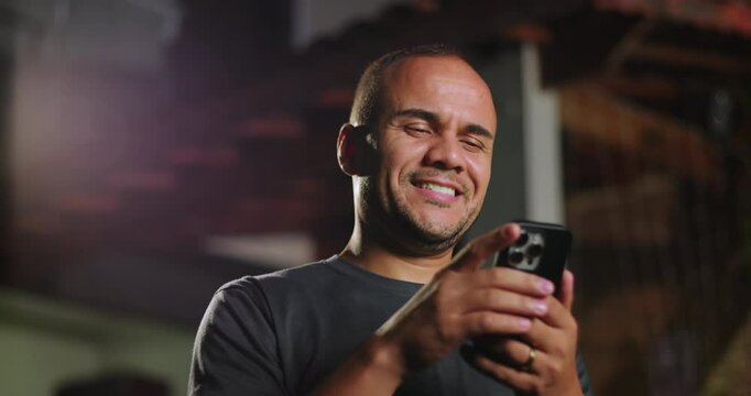 Latin Hispanic man of African descent using smartphone outdoors with slight surprise, reacting to online content while holding phone with both hands in evening setting