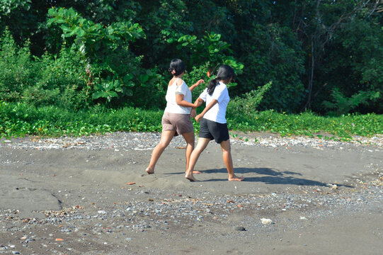 Two teenage girls jog lightly along the beach, smiling and enjoying the morning freshness amid the scenic coastal greenery and soft sandy terrain.