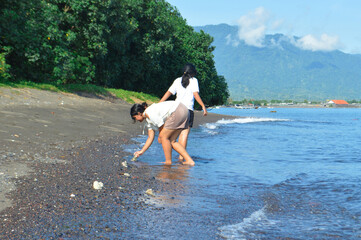 Two girls crouch at the wet sandy shore, closely inspecting something curious in the surf under warm morning light on a tropical beach.