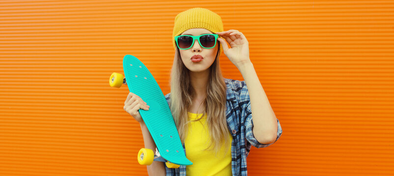 Stylish young woman with skateboard in hat, happy modern teenage girl posing on orange wall