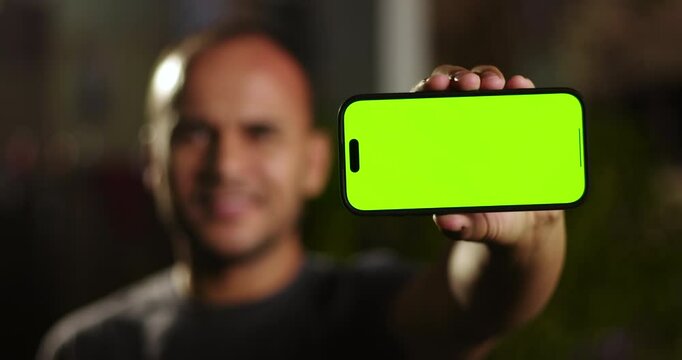 Smartphone with green screen held horizontally by Latin Hispanic man outdoors, close to camera with man blurred in the background
