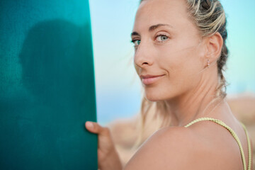Smiling surfer woman with tied blonde ponytail and light gray eyes holds her surfboard while...