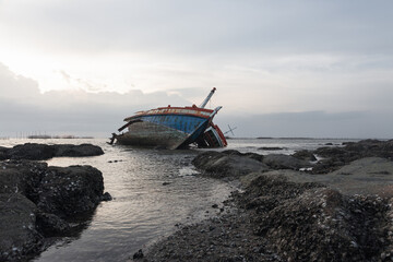 Weathered vessel rests at angle on rugged, rocky shoreline beneath dramatic, moody sky. The scene...