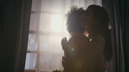 Medium close-up with lens flare of young female servicewoman in khaki military t-shirt hugging beloved biracial teenage son by window in bright morning sun, before leaving for army duty