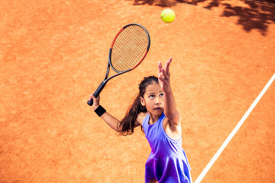 Young girl serving tennis ball on clay court during match