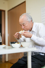 In a modest Japanese apartment, a man in his late 70s wearing a white shirt sits at a small table, eating a bowl of natto rice for breakfast as soft morning light filters gently through the window.