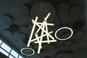 Modern ceiling light fixture with tubular and ring LED lights arranged in a geometric pattern, set against a ceiling with circular acoustic panels and large windows in an industrial-style interior.

