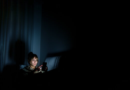 young woman staring at cell phone with face illuminated by screen late at night on sofa