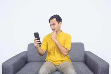 Obraz premium Shocked Asian man wear yellow shirt sit on grey sofa while holding smartphone, Surprised indonesian