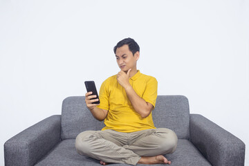 Confused Asian man wear yellow shirt sit on grey sofa holding a phone thinking and choosing isolated