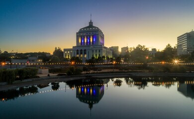Alameda County Courthouse in Oakland, California, USA, is illuminated with blue and yellow lights, reflected in the water at twilight.