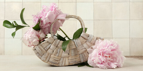 Wicker bag and beautiful peony flowers on table near beige tile wall, closeup