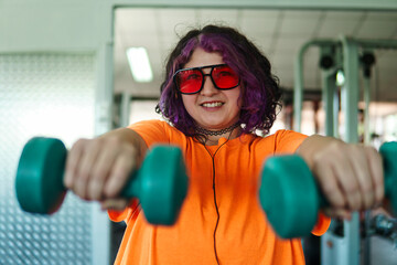 Beautiful young overweight Latina posing and exercising with weights and listening to music, wearing retro glasses and a Discman in a retro-style gym, working hard, very happy. Health and improvement.