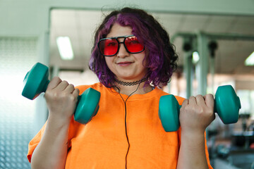 Beautiful young overweight Latina posing and exercising with weights and listening to music, wearing retro glasses and a Discman in a retro-style gym, working hard, very happy. Health and improvement.