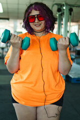 Beautiful young overweight Latina posing and exercising with weights and listening to music, wearing retro glasses and a Discman in a retro-style gym, working hard, very happy. Health and improvement.