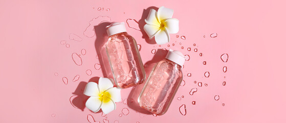Bottles of micellar water, plumeria flowers and water drops on pink background, closeup