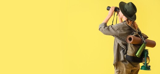 Young female tourist with binoculars and backpack on yellow background with space for text