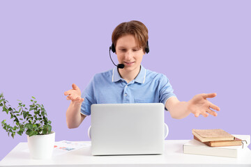 Teenage boy with laptop and headset working at his comfortable workspace on lilac background. Freelance concept