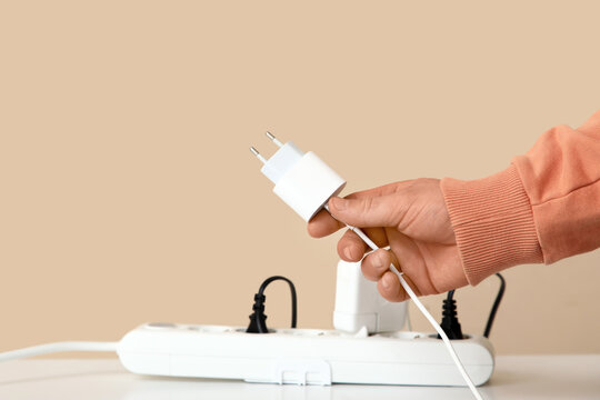 Male hand with unplugged charger and extension cord on table near beige wall, closeup