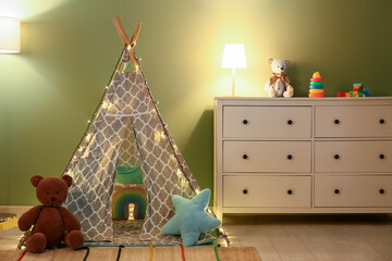 Interior of night children's room with glowing lamps, play tent and toys on commode near green wall