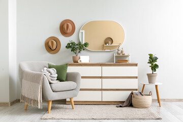 Interior of living room with armchair, hanging mirror, houseplants and home decor on commode near...