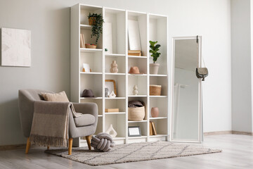 Interior of living room with armchair, mirror, houseplants and home decor in shelving units near white wall