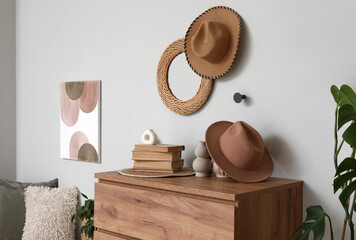 Interior of living room with hanging mirror, houseplants and home decor on commode near grey wall