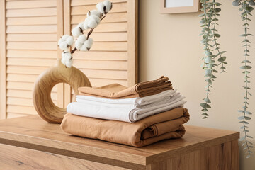 Stack of clean bed sheets with cotton flowers sprig in vase on commode near beige wall