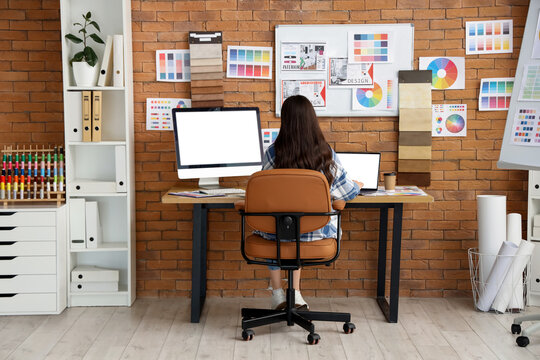 Female designer working with blank computer and laptop at table in office, back view