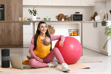 Sporty young woman with her baby in sling and fitball sitting at home