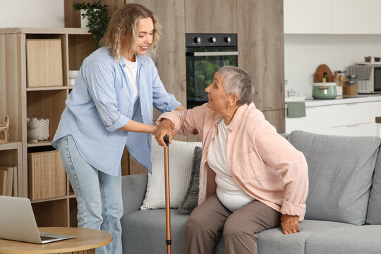 Young woman helping her grandmother with stick to stand up from sofa at home