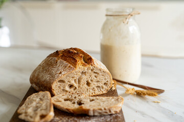 Crispy sourdough bread sits beside a glass jar filled with active starter, accompanied by a rustic cloth and a wooden board in a warm kitchen atmosphere.