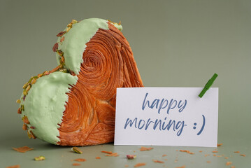 HAPPY MORNING caption on paper note love symbol Heart shaped pistachio croissant on green...